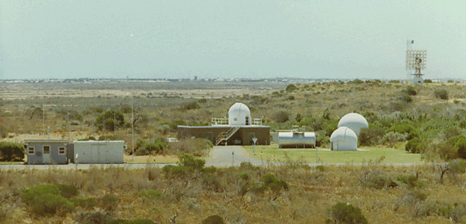 SPAN Site at the height of the Carnarvon Location Project: behind the Security Hut (on the left) are the Riometer antenna back-plane masts; centre is the SPAN building with Razdow telescope dome on the roof; to the right front is the Satellite Tracking Camera dome; just behind is the Radio Telescope radome; and near distant right, the Mobile Laser sat just to the left of the RARR VHF antenna. The Town of Carnarvon can be seen on the far distant horizon.